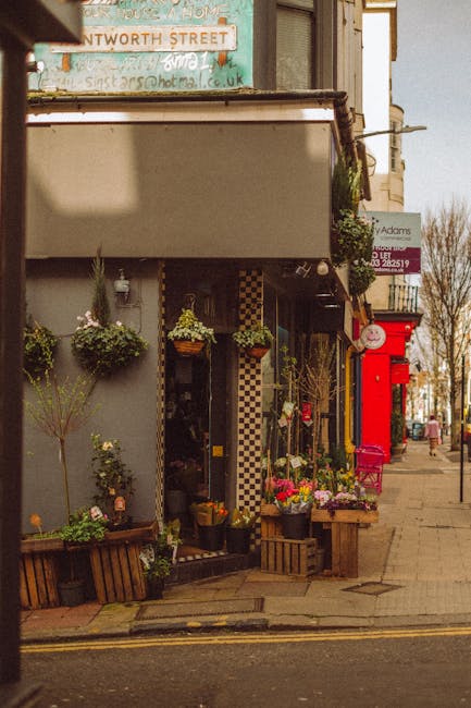 A floral display outside a flower shop on a city street, featuring a bouquet arrangement with fresh pink and purple flowers including roses and carnations, arranged in a clear glass vase. The bouquet 
