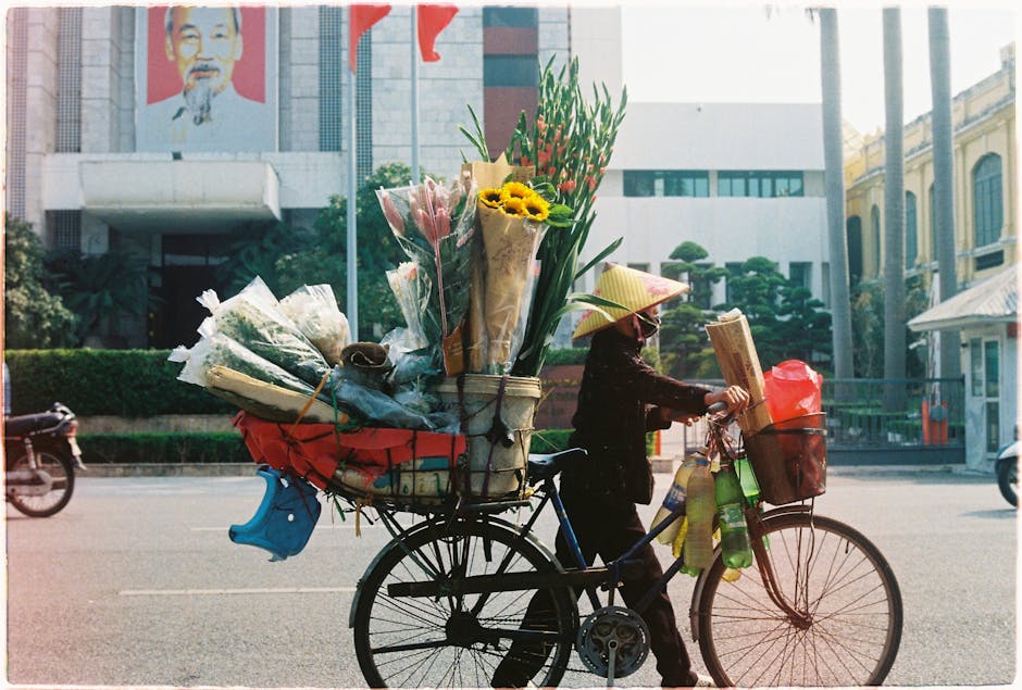 A woman riding a bicycle on a city street carrying an extensive floral arrangement in the rear basket and held in her hands, featuring a variety of freshly cut flowers including sunflowers, pink roses