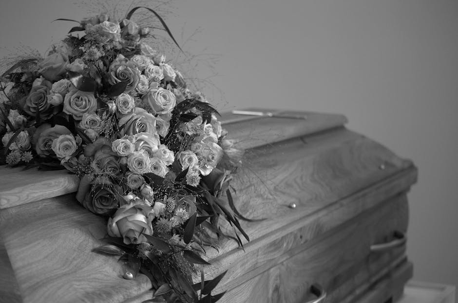 A large, elegant bouquet of fresh flowers rests atop a wooden casket in a funeral setting. The arrangement includes roses in various sizes, with some fully bloomed and others buds, predominantly in li