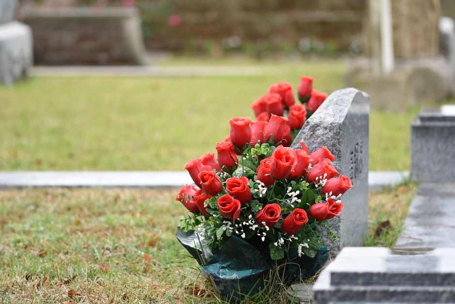 A bouquet of fresh, vibrant red roses with lush green leaves and small white filler flowers, arranged in a dense cluster, rests against a grey gravestone in a cemetery setting. The roses have velvety 